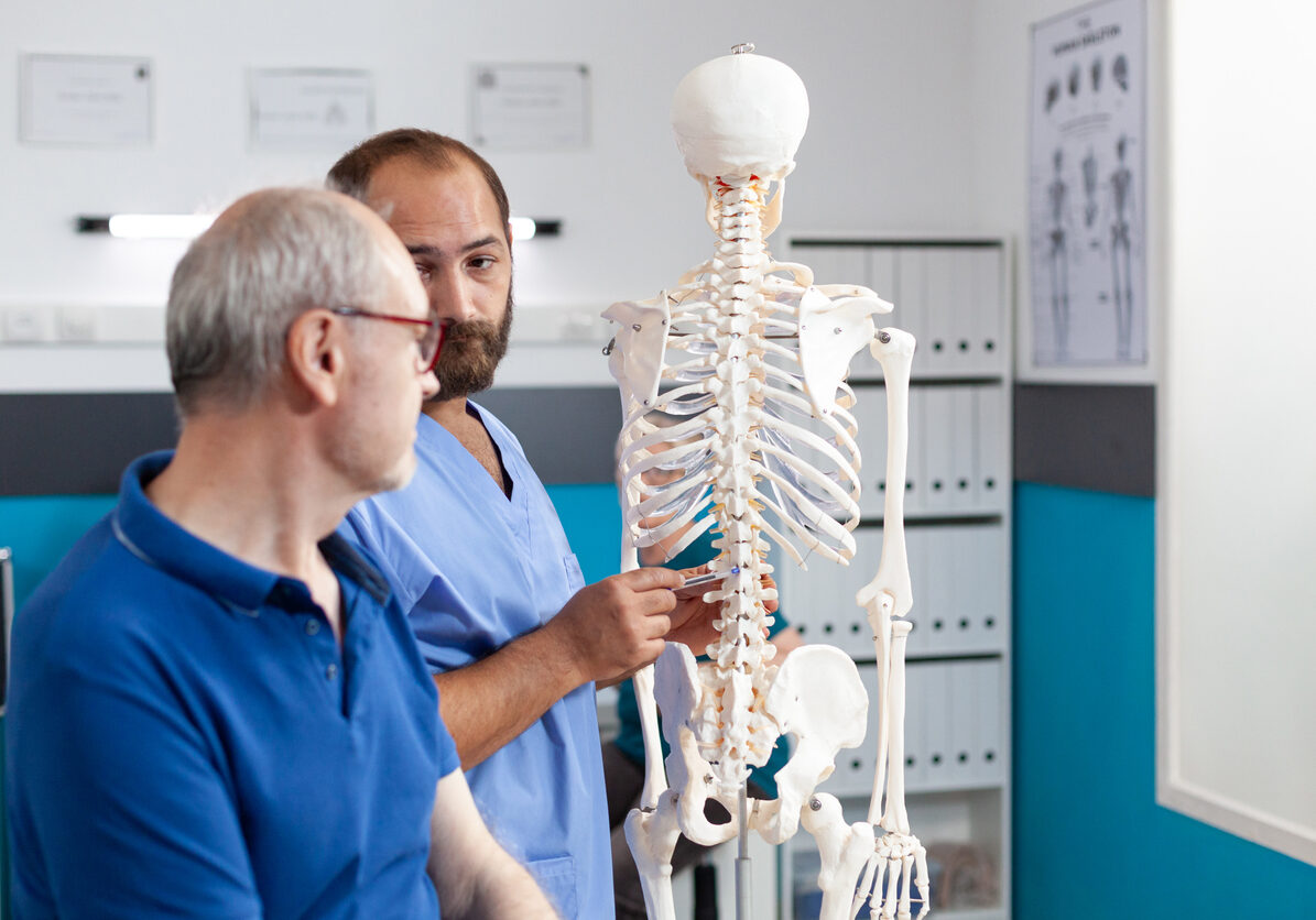 Orthopedist explaining spinal cord on human skeleton to retired patient in physiotherapy office. Chiropractor doing presentation of spine bones and anatomy to old man for osteopathy