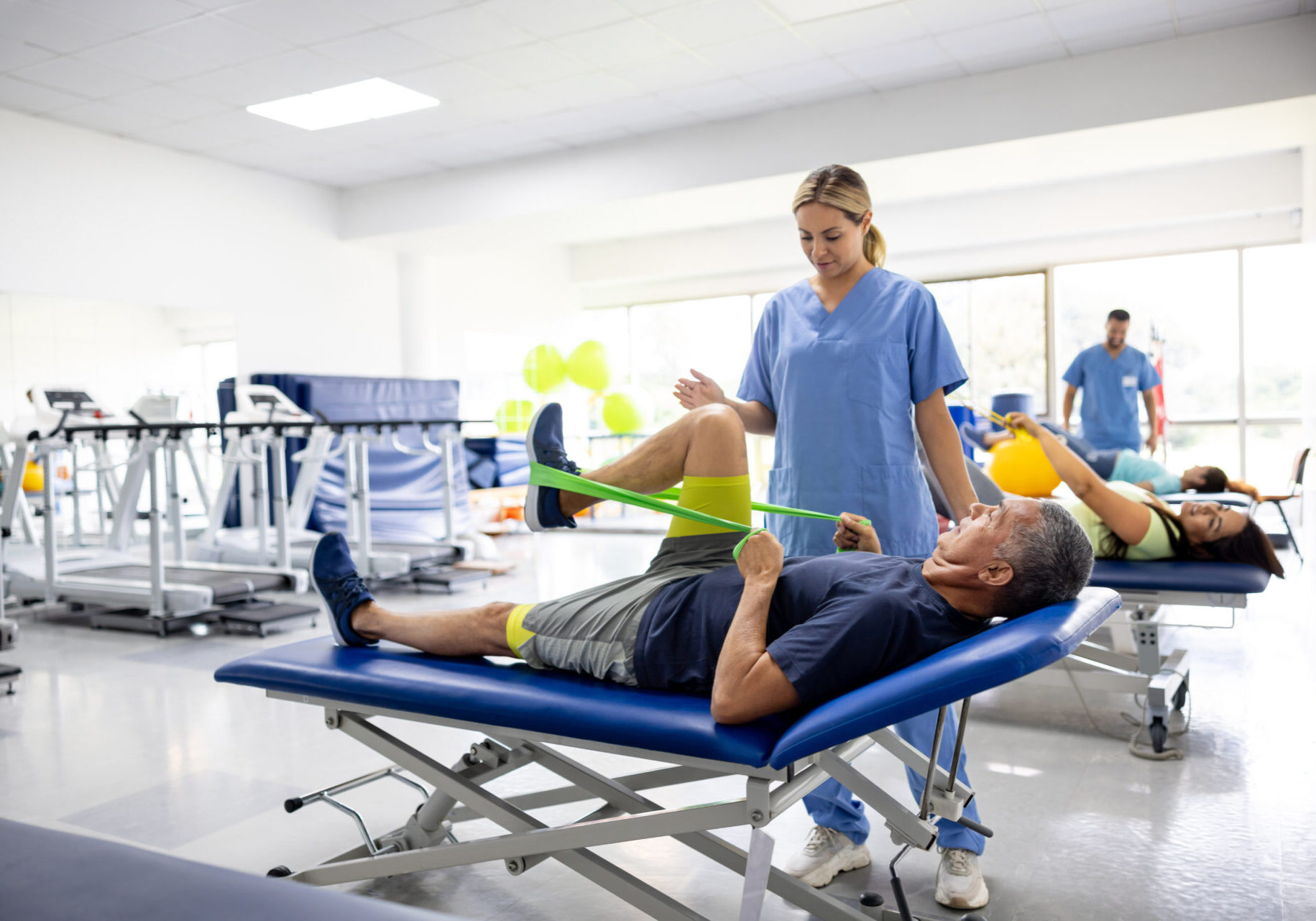 Mature Latin American man doing physical therapy exercises using a stretch band with the assistance of his therapist