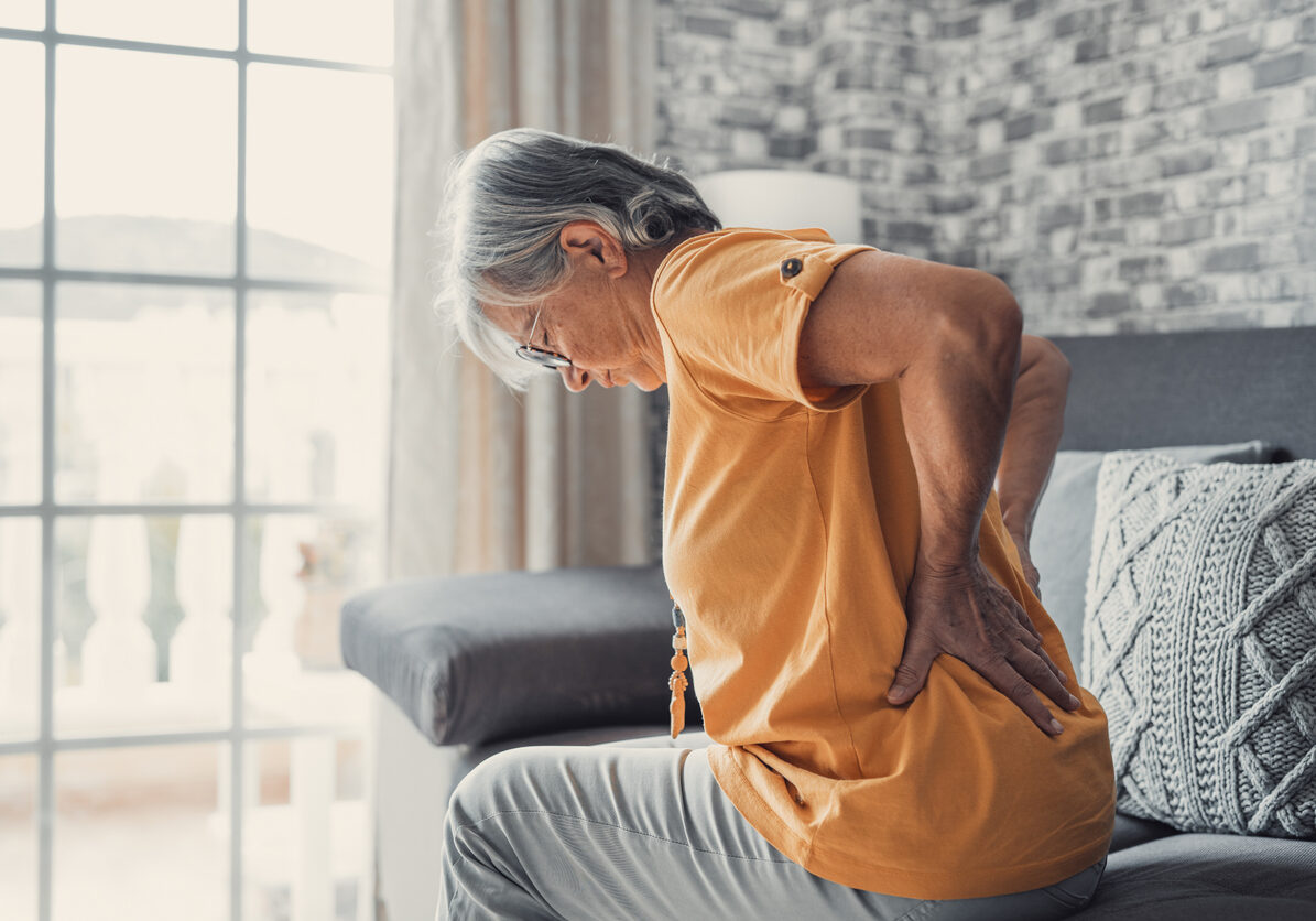 Unhappy mature woman touching back, feeling pain, sitting on couch in living room, unhealthy middle aged senior female suffering from backache, spinal problem, rubbing stiff muscles or kidney