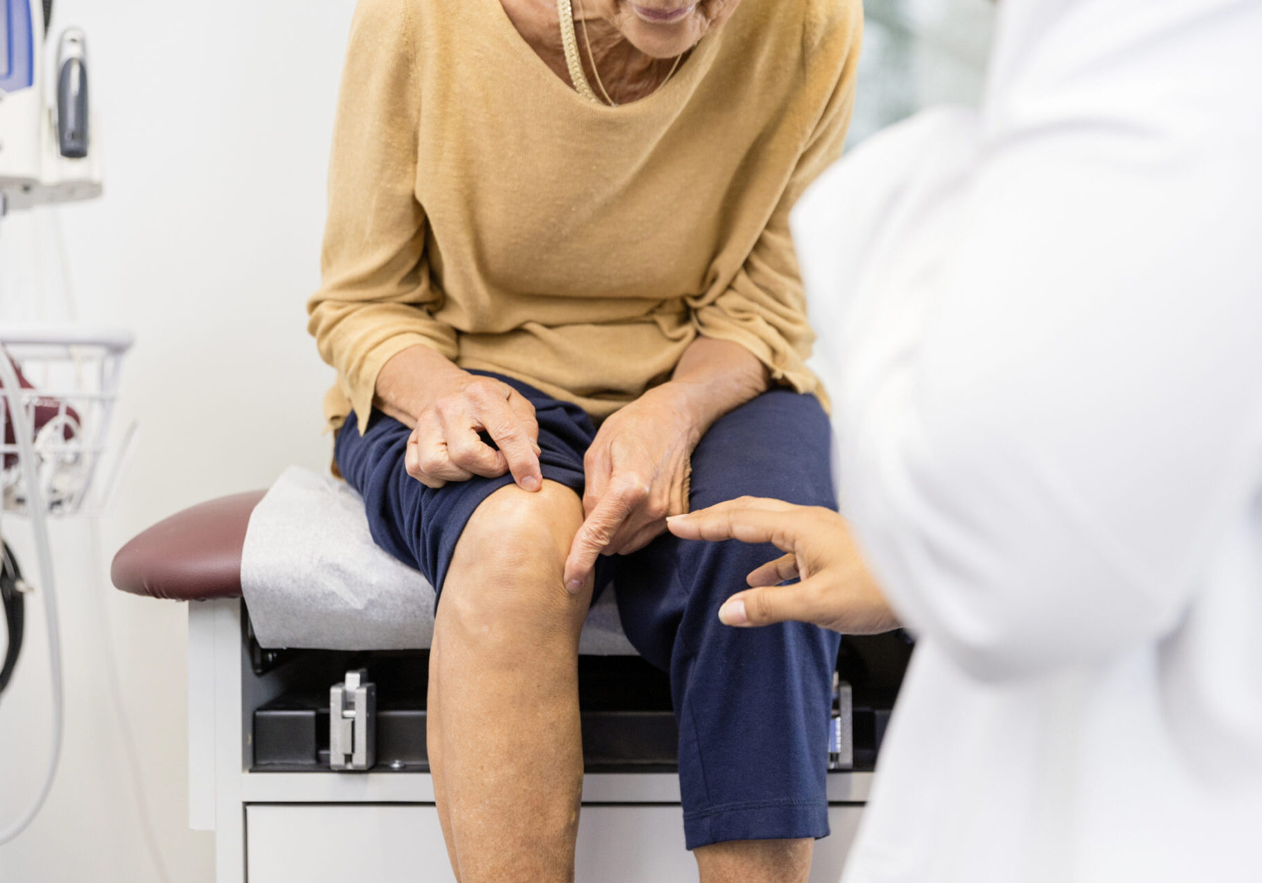 The unrecognizable senior adult woman, sitting on the examination table, shows the unrecognizable female doctor the exact place where her knee hurts.