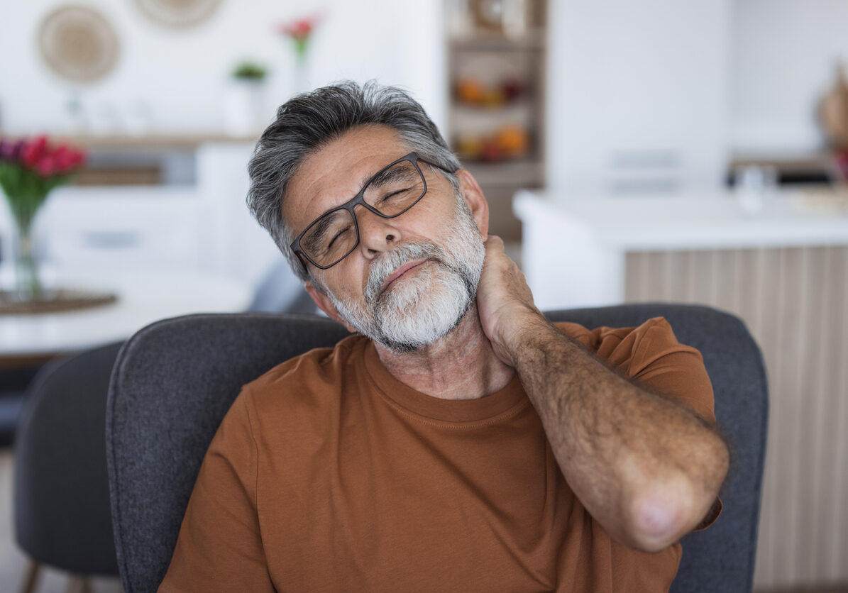Senior man sitting on an armchair at home, suffering from neck pain